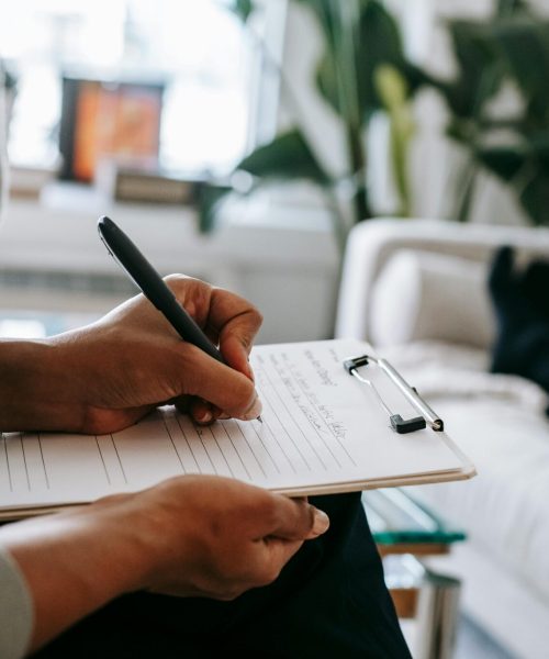 Unrecognizable ethnic female therapist taking notes on clipboard while filling out form during psychological appointment with anonymous client lying on blurred background