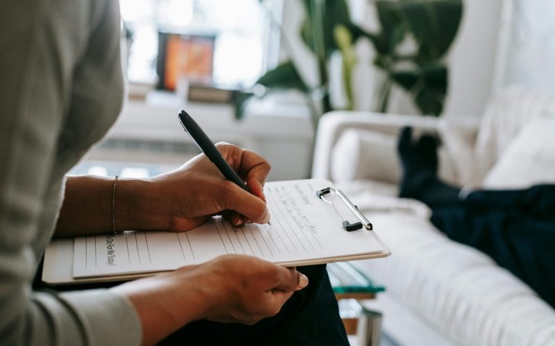 Unrecognizable ethnic female therapist taking notes on clipboard while filling out form during psychological appointment with anonymous client lying on blurred background