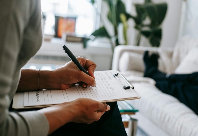 Unrecognizable ethnic female therapist taking notes on clipboard while filling out form during psychological appointment with anonymous client lying on blurred background